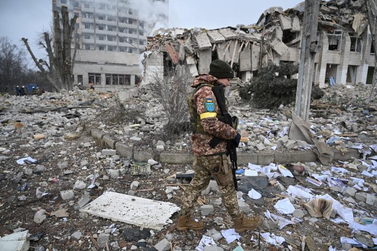 Eine ukrainische Soldatin läuft mit gesenktem Blick und Waffe in der Hand über ein Trümmerfeld.
