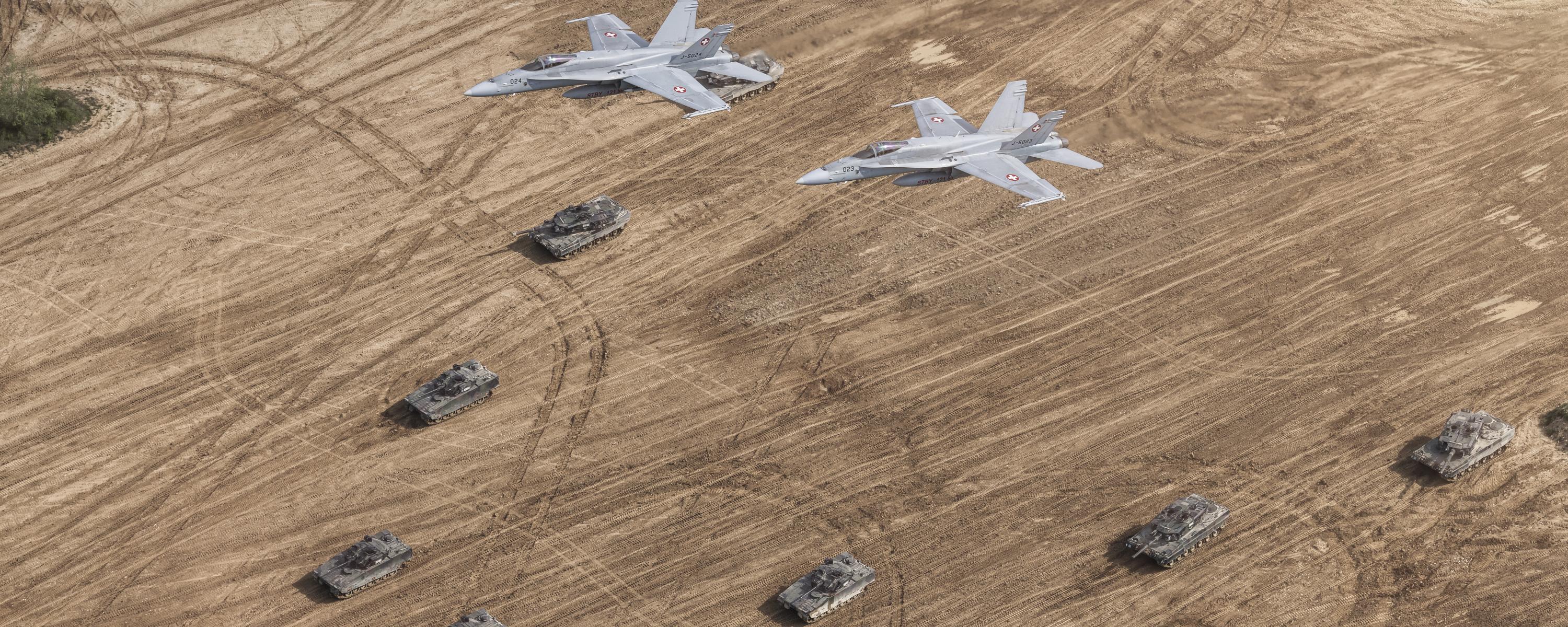 Zwei F/A-18 Kampfjets überfliegen eine Panzer-Keilformation auf einem Acker.