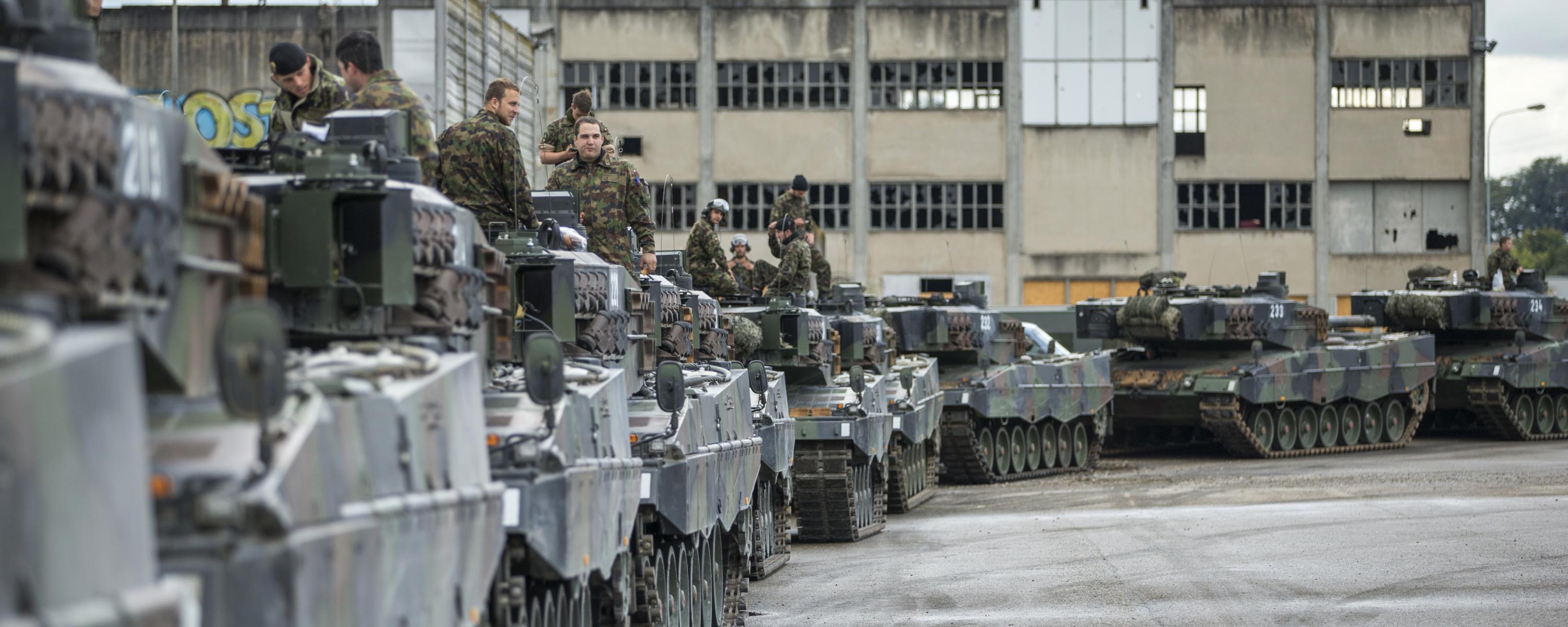 Mehrere Panzer stehen hintereinander auf einem Industriequartier, die Besatzungen sitzen mehrheitlich auf den Panzern.