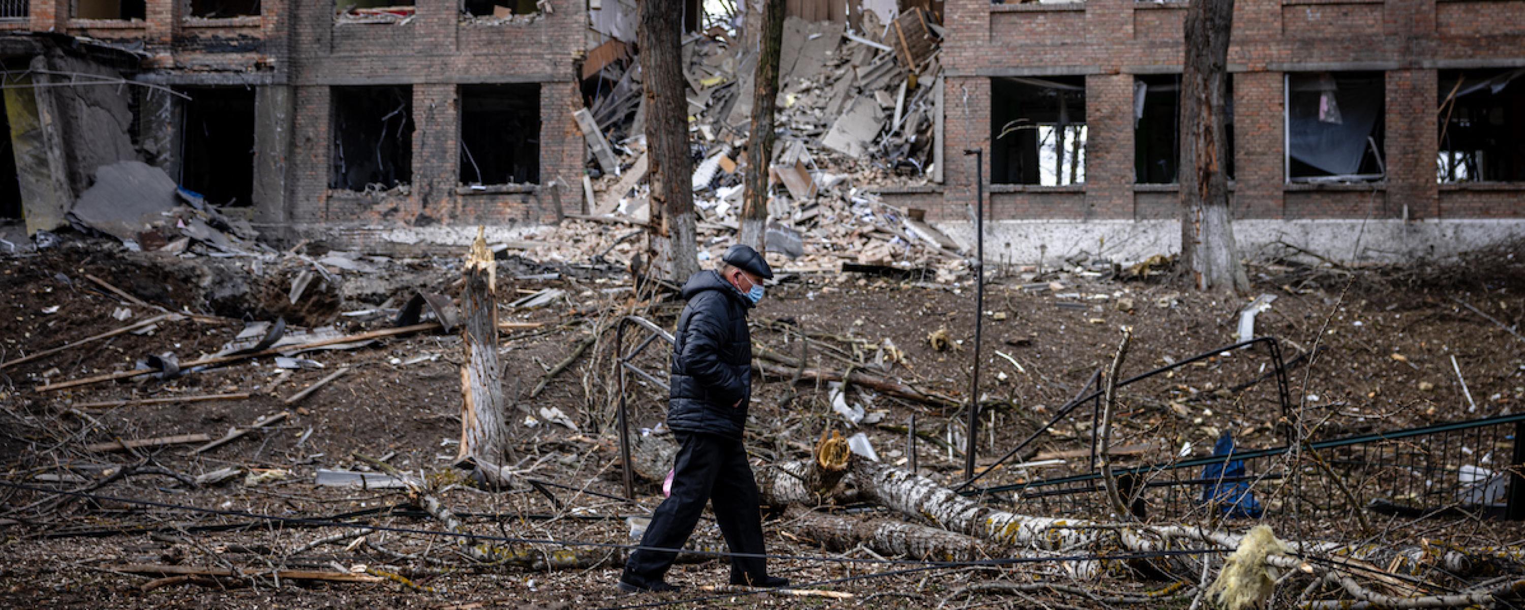 Ein alter Mann geht mit gesenktem Blick durch einen Trümmerhaufen vor einem zerstörten Wohngebäude.