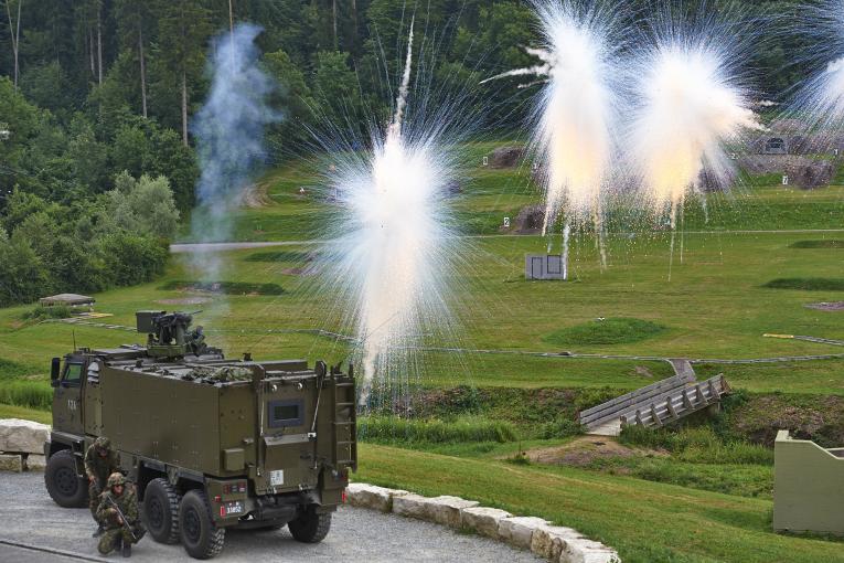 Zwei Soldaten in Kampfmontur suchen Deckung hinter einem GMTF, welches Nebelpetarden schiesst.